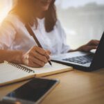 Person writing in notebook using laptop nearby