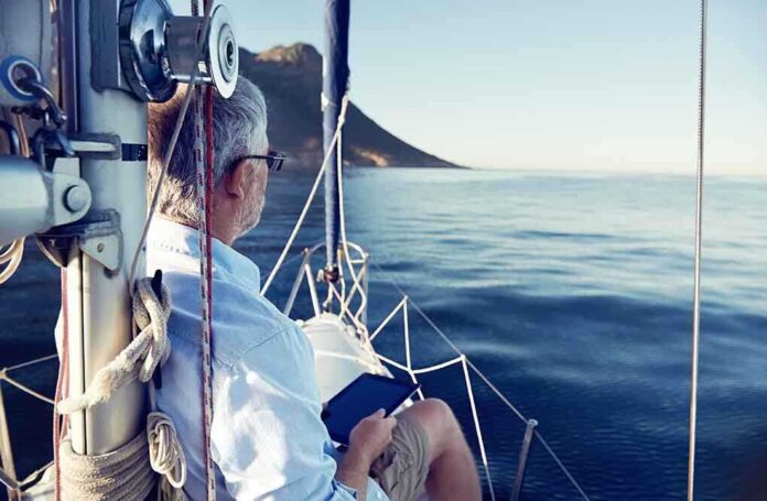 A man sitting on a sailboat, looking out at the calm ocean with mountains in the background