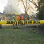 Yellow police tape marking a restricted area at a fire scene with firefighters in the background