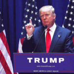 Man speaking at podium with USA flags behind him