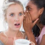 Two women in a casual setting one whispering to the other who looks surprised