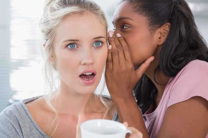 Two women in a casual setting one whispering to the other who looks surprised