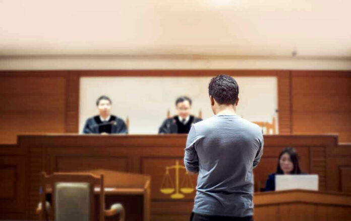 Man stands in courtroom before judges and stenographer.