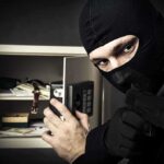A masked burglar holding a gun while attempting to open a safe