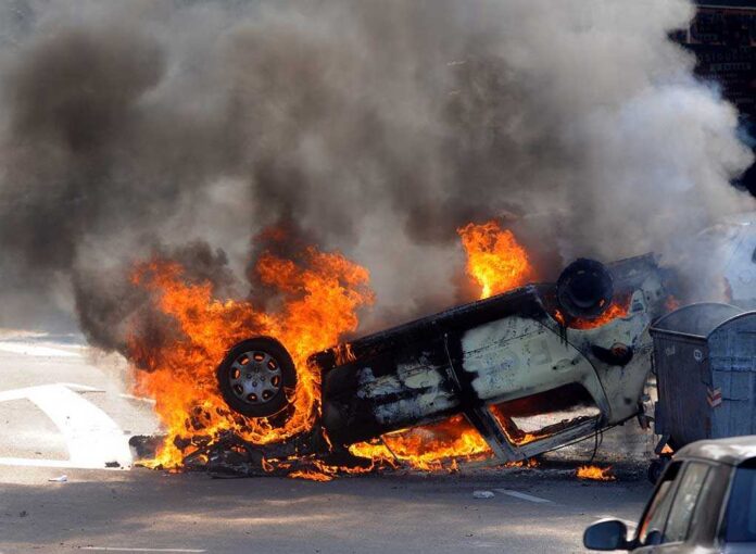 An overturned car engulfed in flames and smoke on a city street