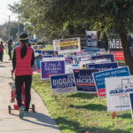 Maniac Socialist PULLS OFF Remarkable Win in Red State Person walking with stroller past numerous election campaign signs.
