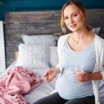 Pregnant woman sitting on bed holding pills and a glass of water