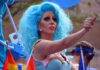 Drag performer with bright blue wig giving thumbs up at parade
