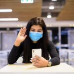 Woman in face mask video calling on smartphone in a cafeteria