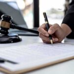 Hand signing legal document beside gavel and laptop
