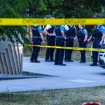 police officers stand behind caution tape at a park scene