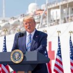 Donald Trump speaking at a podium with American flags and a ship in the background