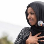 Ilhan Omar speaking at a microphone with hand on chest